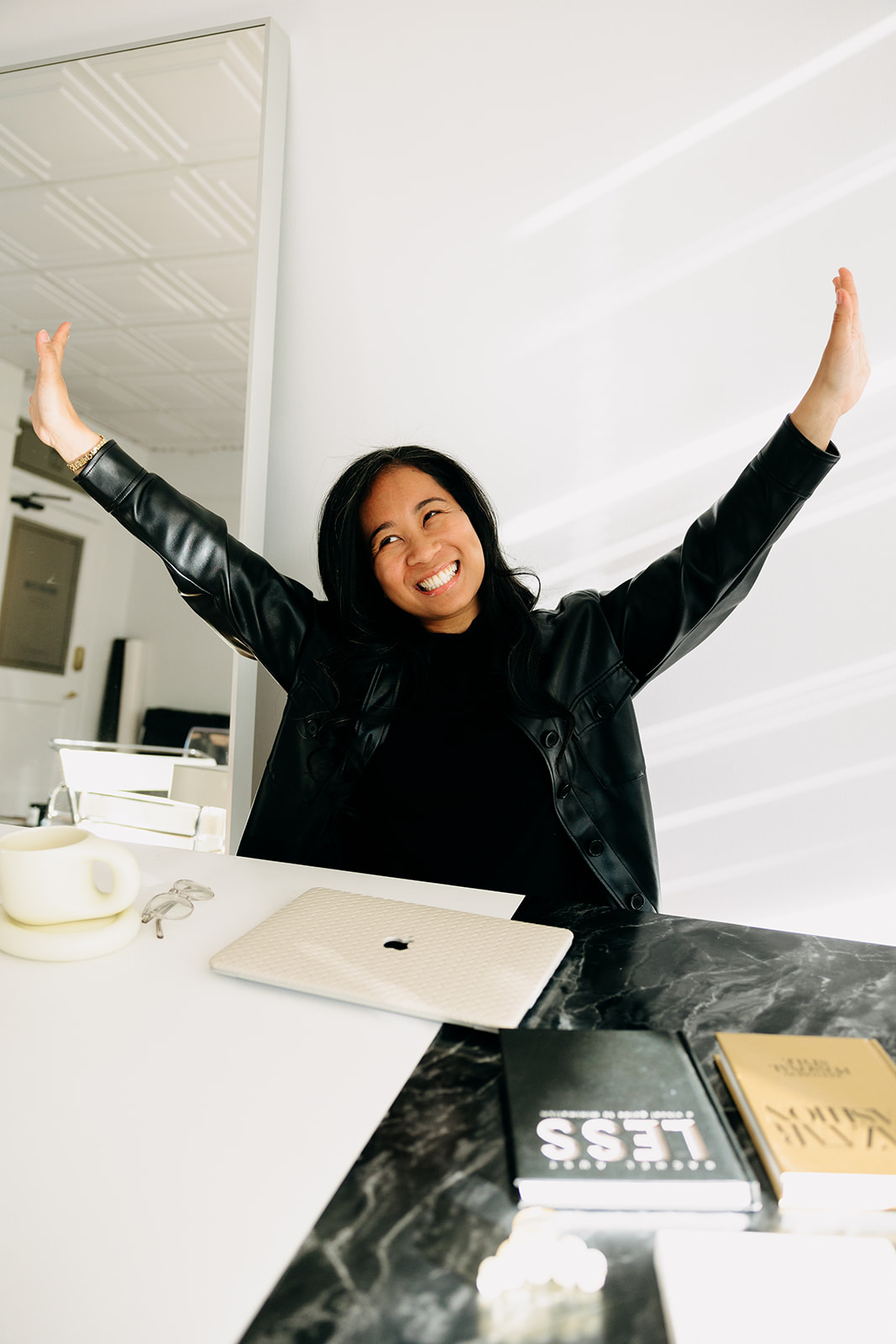 Smiling woman in a leather jacket raising her arms in celebration at her desk — representing a photography mentor confidently launching her wedding photography marketing plan.